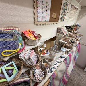 Puede incluir: Un expositor de venta al por menor con varias cestas tejidas llenas de mercanc&iacute;a variada, incluyendo libretas peque&ntilde;as, pegatinas, accesorios para el pelo y bolsitas. Un tablero de corcho con el texto Gabrielle's Gathering est&aacute; montado en la pared.