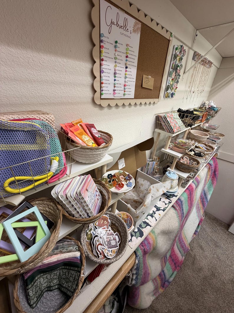 Puede incluir: Un expositor de venta al por menor con varias cestas tejidas llenas de mercanc&iacute;a variada, incluyendo libretas peque&ntilde;as, pegatinas, accesorios para el pelo y bolsitas. Un tablero de corcho con el texto Gabrielle's Gathering est&aacute; montado en la pared.