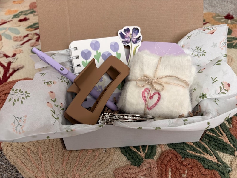 Puede incluir: Una caja de regalo que contiene una pinza para el cabello rectangular de color marr&oacute;n, un cuaderno de espiral con ilustraciones de tulipanes morados, un bol&iacute;grafo morado claro, un cuadrado de tela blanca difusa atado con cordel con un coraz&oacute;n rosa y brazaletes plateados.