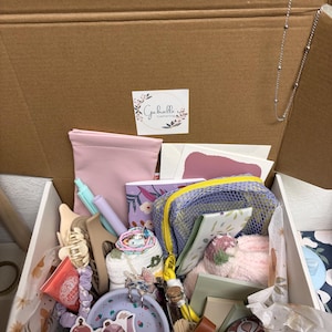 Puede incluir: Una caja de regalo de cart&oacute;n que contiene una colecci&oacute;n de art&iacute;culos que incluye un estuche rosa, una bolsa de malla morada, un cuaderno peque&ntilde;o con flores, pinzas para el pelo, un plato con motivos lavanda, un frasco de vidrio y papeler&iacute;a.
