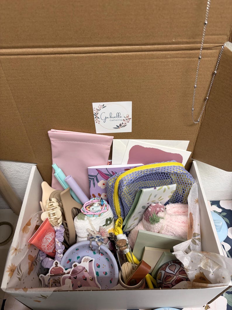 Puede incluir: Una caja de regalo de cart&oacute;n que contiene una colecci&oacute;n de art&iacute;culos que incluye un estuche rosa, una bolsa de malla morada, un cuaderno peque&ntilde;o con flores, pinzas para el pelo, un plato con motivos lavanda, un frasco de vidrio y papeler&iacute;a.