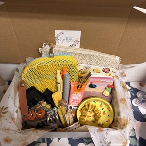 Puede incluir: Una caja de regalo que contiene una bolsa tejida beige, una bolsa de malla amarilla con cremallera, bol&iacute;grafos, pinzas para el cabello, un tubo de crema de manos, una botella de vidrio, un plato de cer&aacute;mica amarillo con naranjas y un coletero amarillo.