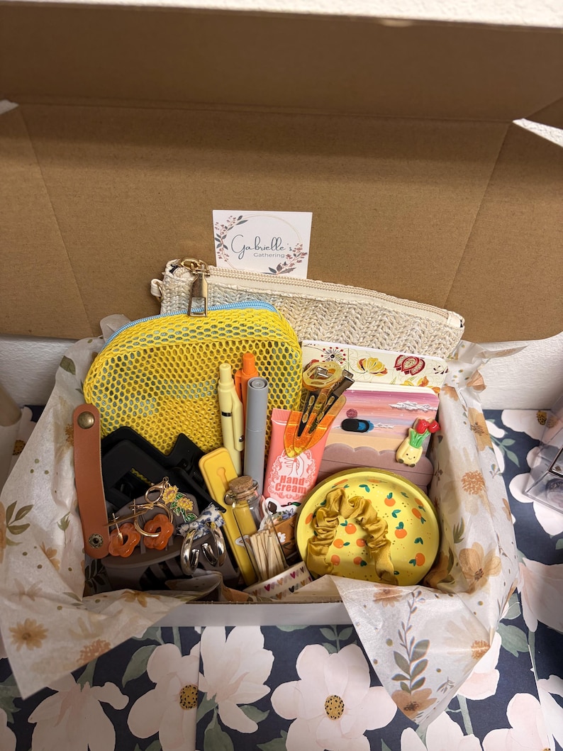 Puede incluir: Una caja de regalo que contiene una bolsa tejida beige, una bolsa de malla amarilla con cremallera, bol&iacute;grafos, pinzas para el cabello, un tubo de crema de manos, una botella de vidrio, un plato de cer&aacute;mica amarillo con naranjas y un coletero amarillo.