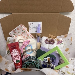 Puede incluir: Una caja de regalo que contiene un tubo de crema de manos de cereza, una botella de vidrio peque&ntilde;a con cerillas, un bol&iacute;grafo morado claro, calcetines blancos difusos, pendientes de aro morados con forma de flor y un marco verde.