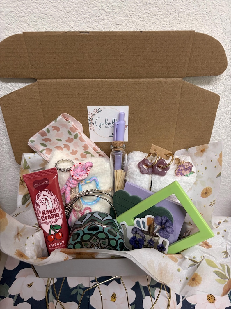 Puede incluir: Una caja de regalo que contiene un tubo de crema de manos de cereza, una botella de vidrio peque&ntilde;a con cerillas, un bol&iacute;grafo morado claro, calcetines blancos difusos, pendientes de aro morados con forma de flor y un marco verde.