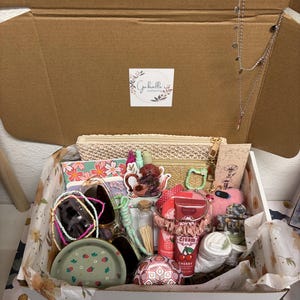 Puede incluir: Una caja de regalo de cart&oacute;n que contiene un estuche tejido beige, un cuaderno floral, un plato verde con estampado de fresas, un collar plateado, crema de cereza, un coletero rosa, un frasco de vidrio con cerillas y varios accesorios.