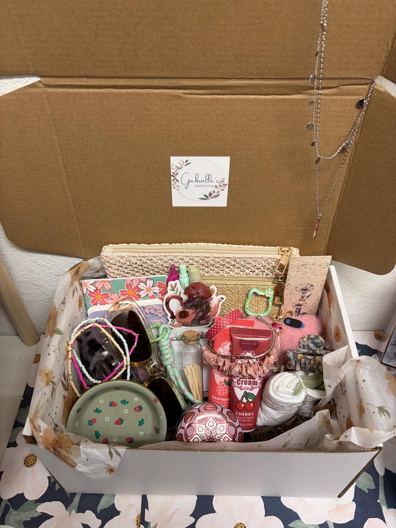 Puede incluir: Una caja de regalo de cart&oacute;n que contiene un estuche tejido beige, un cuaderno floral, un plato verde con estampado de fresas, un collar plateado, crema de cereza, un coletero rosa, un frasco de vidrio con cerillas y varios accesorios.