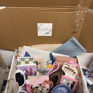 Puede incluir: Una caja de regalo de cart&oacute;n llena de art&iacute;culos variados, incluyendo gafas de sol con montura blanca, b&aacute;lsamo labial rosa, un plato azul de cer&aacute;mica con un pato, un marco de fotos rosa, accesorios para el cabello y un collar de plata.