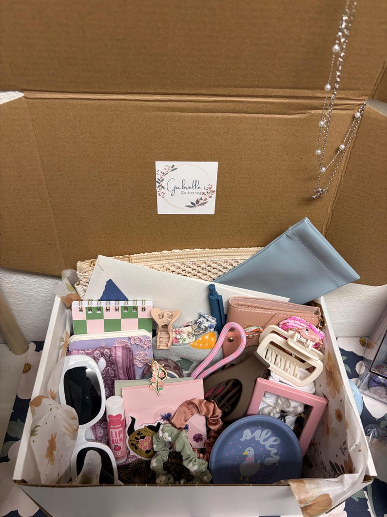 Puede incluir: Una caja de regalo de cart&oacute;n llena de art&iacute;culos variados, incluyendo gafas de sol con montura blanca, b&aacute;lsamo labial rosa, un plato azul de cer&aacute;mica con un pato, un marco de fotos rosa, accesorios para el cabello y un collar de plata.