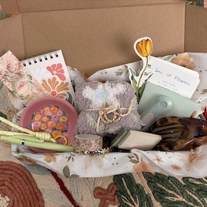 Puede incluir: Una caja de regalo de cart&oacute;n que contiene un cuaderno de espiral con dise&ntilde;os florales, un plato rosa con motivos de flores, un par de calcetines suaves de color malva, un bol&iacute;grafo verde claro, un rollo de cinta estampada, una pegatina de tulip&aacute;n, un bloc de notas con el texto Sea of Flowers y una pinza para el pelo.