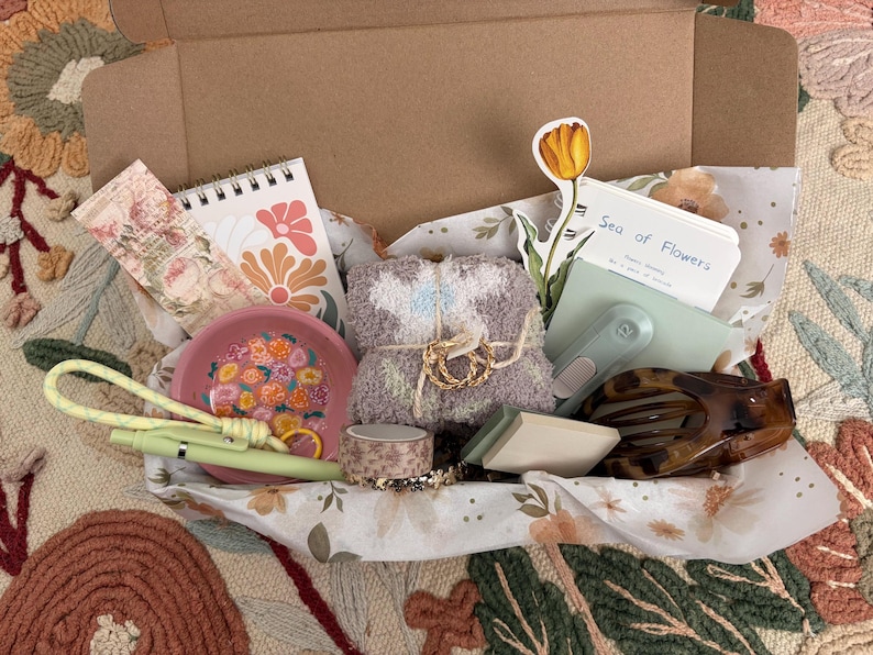 Puede incluir: Una caja de regalo de cart&oacute;n que contiene un cuaderno de espiral con dise&ntilde;os florales, un plato rosa con motivos de flores, un par de calcetines suaves de color malva, un bol&iacute;grafo verde claro, un rollo de cinta estampada, una pegatina de tulip&aacute;n, un bloc de notas con el texto Sea of Flowers y una pinza para el pelo.