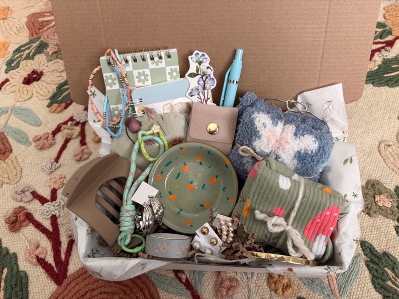 Puede incluir: Una caja de regalo que contiene un plato de cer&aacute;mica verde con detalles en naranja, un bol&iacute;grafo azul claro, una cartera beige, una bolsa azul difusa, un art&iacute;culo de tela acanalada verde, una pinza para el cabello, joyas de cuentas, un cuaderno peque&ntilde;o, una pegatina floral y varios pendientes.