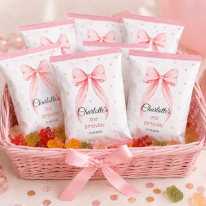 Op de afbeelding: Een lichtroze gevlochten mand met diverse witte snackzakjes versierd met roze stippen en een grote roze strikillustratie. Elk zakje bevat de tekst Charlotte's 2nd Birthday 17.05.2026. De mand is gestrikt met een roze lint.