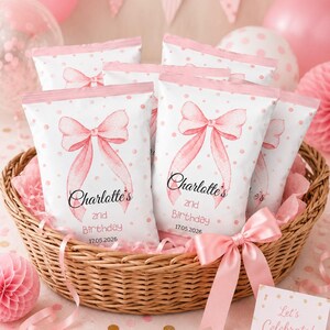 Op de afbeelding: Een rieten mand gevuld met witte zakken met roze stippen en een aquarel strik ontwerp. Elke zak is voorzien van de tekst "Charlotte's 2nd Birthday 17.05.2026". Roze decoraties en een kaartje "Let's Celebrate!" maken de scène compleet.
