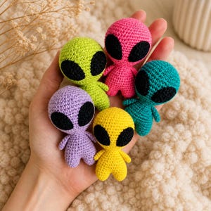 Könnte beinhalten: Fünf kleine, gehäkelte Alienfiguren in Lila, Grün, Pink, Gelb und Türkis. Jede Figur hat große, schwarze, ovale Augen und kleine Arme und Beine. Die Aliens werden in einer Hand gehalten.