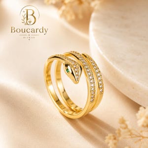 May include: A gold-toned snake ring featuring a coiled design with a row of small clear stones along the band and a single green stone for the eye. The brand name Boucardy Bijoux is printed in the upper left corner with a logo of a letter B.