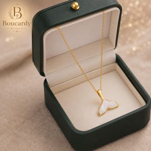 Può includere: Una collana a catena dorata con un ciondolo a forma di coda di balena bianca all'interno di un portagioie verde scuro. Il coperchio della scatola presenta il testo Boucardy Bijoux con un logo. Il ciondolo ha una superficie bianca curva.