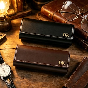 Puede incluir: Dos carteras de llaves de cuero, una negra y otra marrón, se exhiben sobre una superficie de madera. Cada cartera tiene costuras blancas y las iniciales doradas "DK". También se ven un reloj de pulsera y gafas.