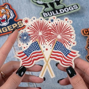 May include: Embroidered patches featuring patriotic designs. One patch showcases crossed American flags with fireworks, while others display a tiger, bulldog, and the word "Bulldogs". The patches are in various colors and styles.