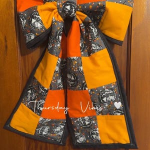 Puede incluir: Un gran lazo decorativo con un diseño de patchwork de tela naranja sólida y tela gris estampada con calabazas de Halloween en blanco y negro. El lazo tiene un borde negro y el texto "Thursday Vibes" con un corazón blanco al frente.