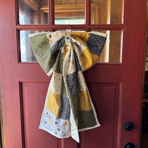 May include: A decorative fabric bow featuring a patchwork design with various patterns in shades of yellow, black, olive green, and white. The fabric displays motifs of honeybees, honeycomb structures, and small floral or leaf-like prints.