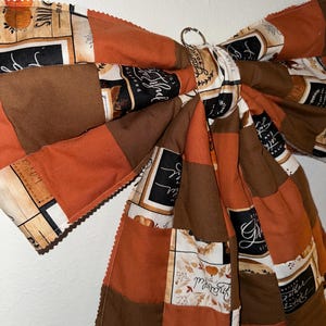 Op de afbeelding: Een grote stoffen strik met een patchworkontwerp in tinten oranje, bruin en crème. De stof toont gedrukte tekst, waaronder de woorden Grateful, Blessed en Gather, naast illustraties van pompoenen en herfstbladeren.