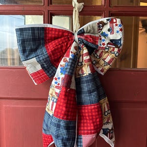 May include: A large, decorative bow made of patchwork fabric, featuring red, white, and blue patterns. The bow is suspended by a cream-colored lace ribbon. The fabric includes plaid, stars, and patriotic motifs, suitable for holiday or seasonal decor.