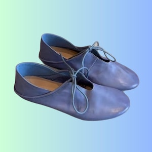 Può includere: Un paio di ballerine in pelle azzurra con lacci coordinati. Le scarpe hanno una punta arrotondata e un profilo basso, adatte per un abbigliamento casual. I lacci sono annodati con un fiocco sulla parte superiore del piede. Lo sfondo ha una sfumatura di blu e verde.