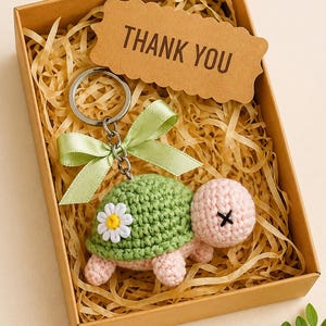 May include: A crochet turtle keyring with a green shell and pink body, decorated with a white daisy. It's presented in a brown gift box with a "THANK YOU" tag and green ribbon.