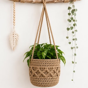 May include: A tan crochet hanging planter basket with a textured diamond pattern, suspended by two matching cords from a horizontal piece of driftwood. The driftwood is decorated with a hanging wooden bead tassel and a trailing green vine plant.