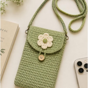 May include: A sage green crochet crossbody bag featuring a long strap, a flap closure adorned with a white and green crocheted flower, and a single light brown wooden button. The bag is constructed with a dense, uniform stitch pattern.