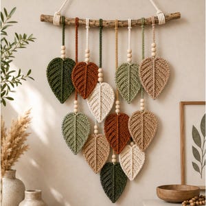 May include: A wall hanging featuring multiple crocheted leaf shapes in shades of dark green, rust, sage, cream, and tan. The leaves are suspended from a natural wooden branch by cotton cords accented with small, round, light-colored wooden beads.