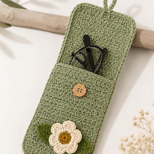 May include: A sage green crocheted glasses case featuring a small wooden button closure and a decorative white flower appliqué with two dark green leaves. The case has a top loop for hanging and contains a pair of dark-rimmed spectacles.