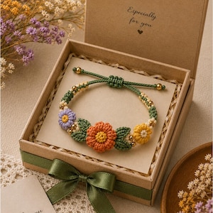 May include: A floral macrame bracelet with orange, yellow, and purple flowers, presented in a gift box with a green ribbon. The box is open, revealing the bracelet on a card that says "Especially for you."