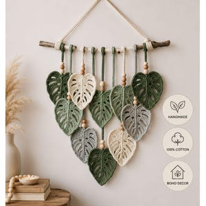 Könnte beinhalten: Ein handgefertigter Boho-Wandschmuck mit gehäkelten Monstera-Blättern in Grün-, Grau- und Cremetönen, aufgehängt an einem natürlichen Ast. Die Blätter sind mit Holzperlen und einer Baumwollschnur verziert. Der Text "HANDMADE", "100% COTTON" und "BOHO DECOR" ist enthalten.