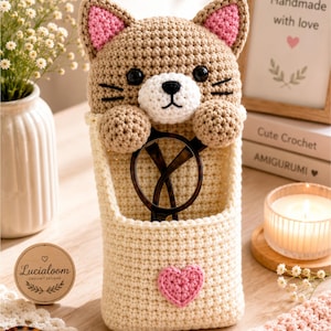 Può includere: Un portaocchiali all'uncinetto a forma di gatto beige con orecchie interne rosa e un'applicazione a forma di cuore rosa sul davanti. Il gatto ha occhi neri, un muso bianco e due piccole zampe appoggiate sul bordo superiore.