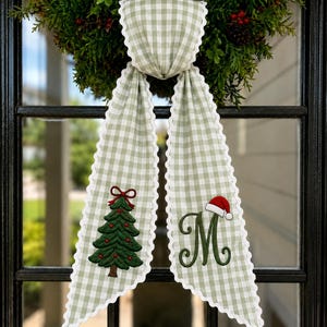 May include: A sage green and white gingham fabric ribbon with scalloped white edges. The left tail features an embroidered green Christmas tree with a red bow, and the right tail features an embroidered green letter M with a red Santa hat.