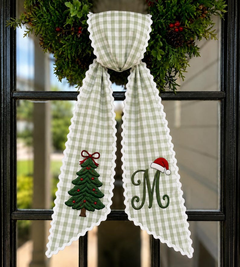 May include: A sage green and white gingham fabric ribbon with scalloped white edges. The left tail features an embroidered green Christmas tree with a red bow, and the right tail features an embroidered green letter M with a red Santa hat.