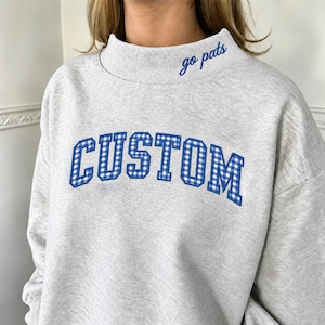 May include: A light gray mock neck sweatshirt featuring the word CUSTOM in blue and white gingham block lettering across the chest. The collar includes the embroidered text go pats in a blue cursive script on the right side of the neck.
