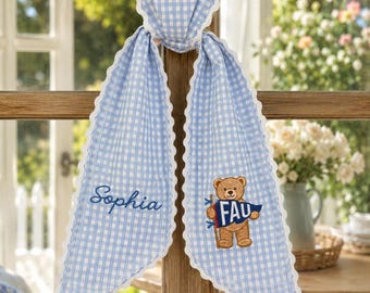 May include: A light blue and white gingham fabric heart ornament with two long, pointed tails featuring white scalloped edges. The left tail is embroidered with the name Sophia in blue, and the right tail features an embroidered brown teddy bear holding a blue and white pennant with the letters FAU.