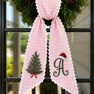 May include: A pink and white gingham fabric ribbon with scalloped white edges. The ribbon features an embroidered green Christmas tree with a red bow on the left tail and a large green letter A wearing a red and white Santa hat on the right tail.