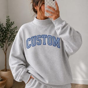May include: A light gray long-sleeve sweatshirt with a mock neck collar. The front features the word CUSTOM in a blue and white gingham check pattern with a dark blue outline, presented in a bold, arched, collegiate-style block font.