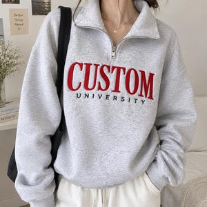 May include: A light gray quarter-zip sweatshirt featuring the word CUSTOM in large red embroidered letters with the word UNIVERSITY in smaller black letters underneath. The garment has a high collar with a silver zipper and long sleeves.