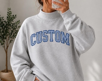May include: A light gray long-sleeve sweatshirt with a mock neck collar. The front features the word CUSTOM in a blue and white gingham check pattern with a dark blue outline, presented in a bold, arched, collegiate-style block font.