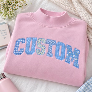 May include: A light pink mock neck sweatshirt featuring the word CUSTOM in large, blue, patterned applique letters. Each letter displays a different design, including gingham, polka dots, stripes, and floral prints, stitched onto the fabric.