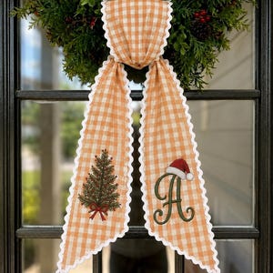 May include: A long orange and white gingham ribbon with scalloped white edges. The left side features an embroidered green Christmas tree with a red bow, and the right side displays a large green letter A topped with a red and white Santa hat.