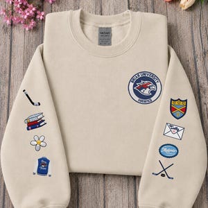 May include: A beige sweatshirt with embroidered patches. The patches include a hockey stick, books, a flower, a varsity jacket, a shield, an envelope, and a logo that says "Malone's". The sweatshirt also has a Briar University Hawks logo.