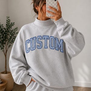 May include: A light gray mock neck sweatshirt featuring the word CUSTOM in blue and white gingham patterned block letters across the chest. The sweatshirt has long sleeves with ribbed cuffs and a matching ribbed hem, paired with light gray sweatpants.