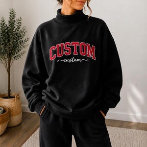 May include: A black long-sleeve sweatshirt with a mock neck collar. The front features the word CUSTOM in large red block letters with a white outline, and the word custom written below it in a white cursive script font.