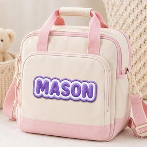 May include: A cream-colored lunch bag with pink trim, pink handles, and a detachable shoulder strap. The front features a rectangular patch with the name MASON in purple bubble letters with a white outline. It has a zippered front pocket.